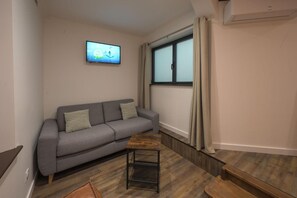 Uma sala de estar moderna com um sofá cinza, uma pequena mesa de centro de madeira, uma televisão montada na parede e uma janela com cortinas.