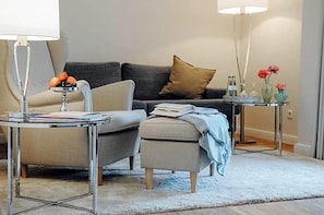 Una sala moderna con un sofá gris, una mesita redonda con una lámpara y una mesita auxiliar con objetos decorativos.