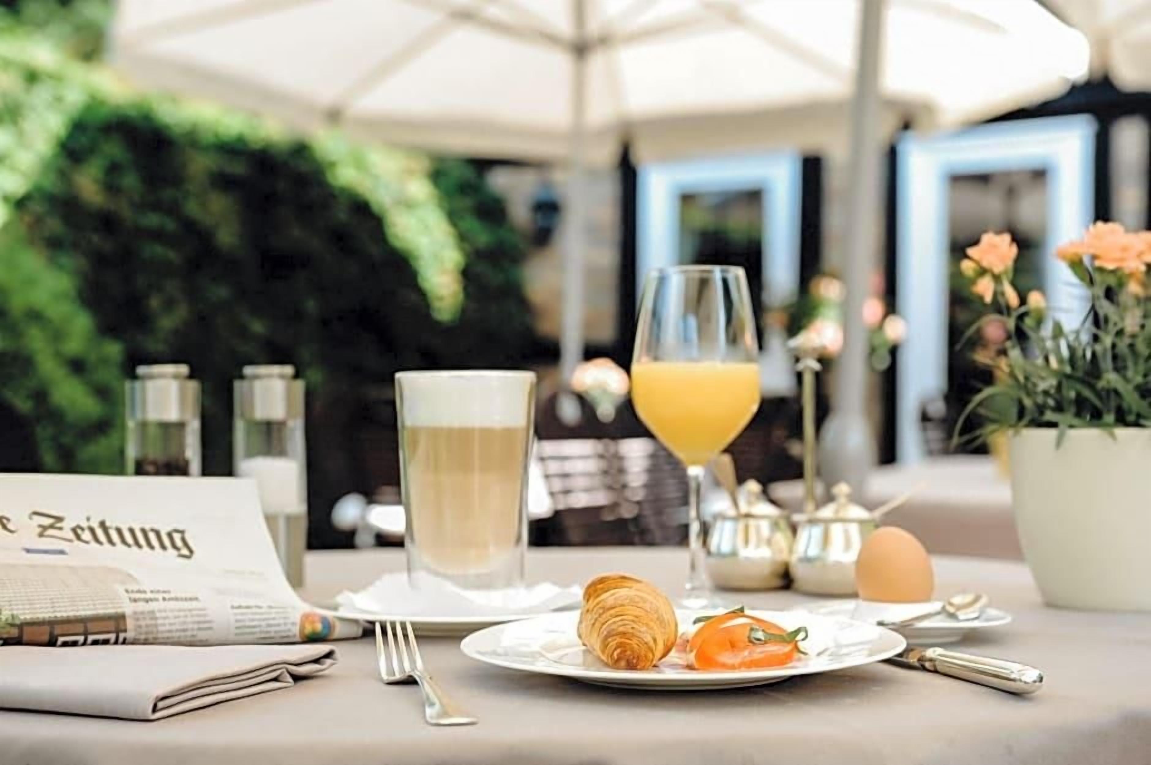 A breakfast setting with a croissant, a glass of orange juice, a newspaper, and a glass of white beverage, likely milk, on a table with a menu.