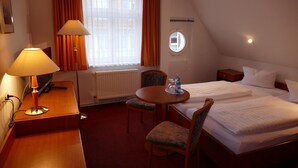 A hotel room with two beds, a desk with a lamp, a chair, a small table with a bottle, and a window with curtains. - Hotel Am Dom (Greifswald)