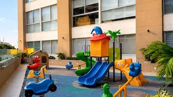 Un parc infantil amb diverses estructures de joc, com tobogans, cadenes i un espai per escalar, al costat d'un edifici amb nombroses finestres.