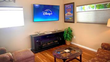 Een woonkamer met een flatscreen-tv waarop het Disney+-logo te zien is, een ingelijst reisposter, een salontafel met glazen blad en een open haard.