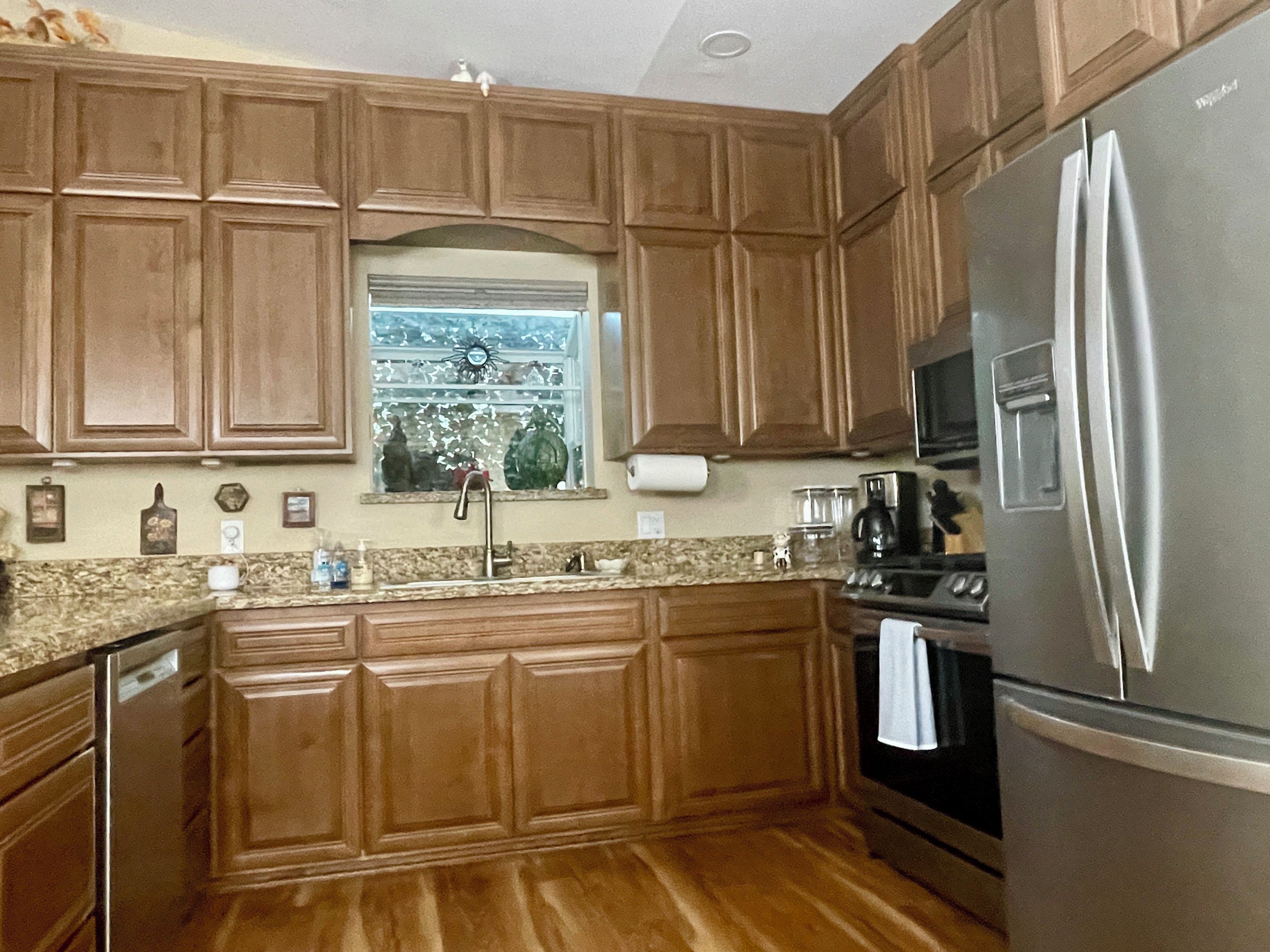 A kitchen with wooden cabinets, a stainless steel refrigerator, a microwave, and a window with a view of trees.