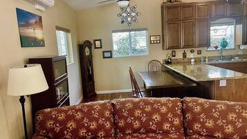 A living room with a sofa, a dining table, and a kitchen area with cabinets and a window.
