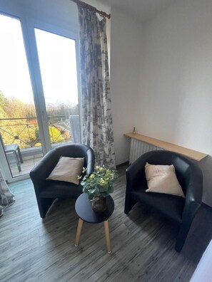 Una habitación con dos sillones negros, una mesita redonda con una planta, vistas a un balcón con barandilla y una ventana con cortinas.