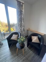 A room with two black armchairs, a small round table with a plant, a view of a balcony with a railing, and a window with curtains.
