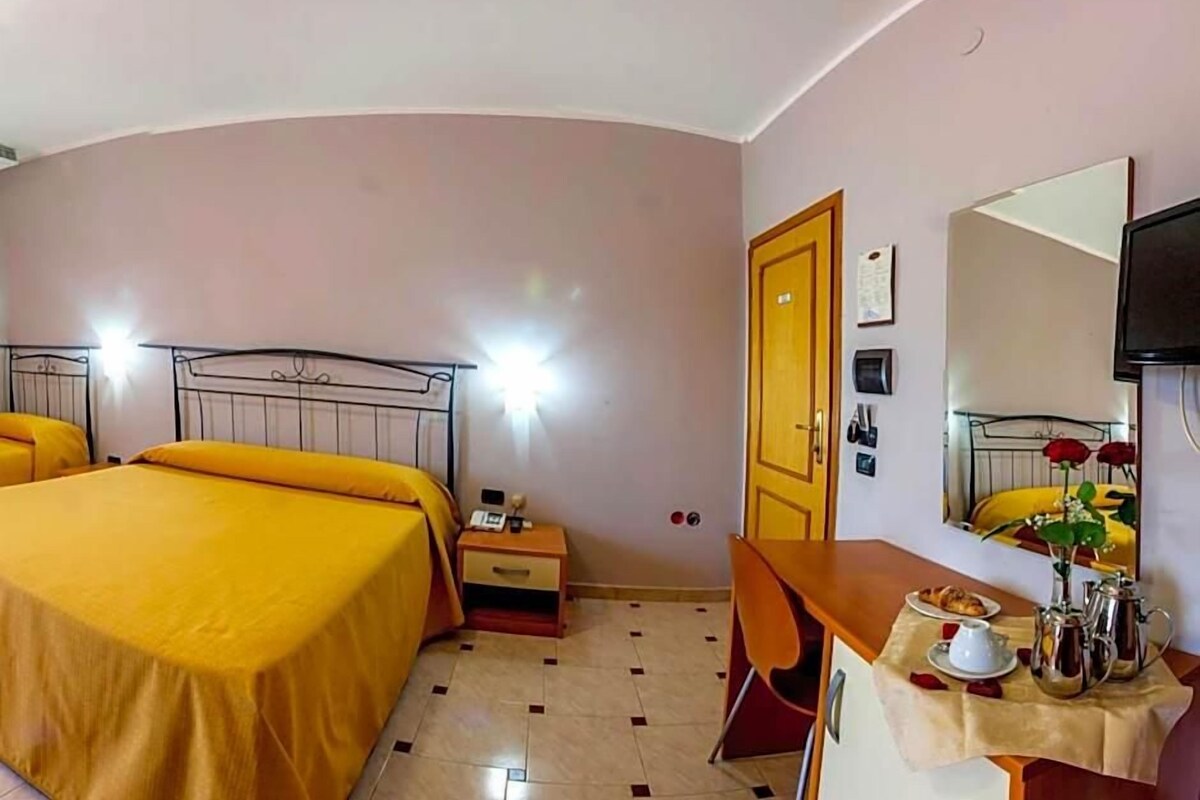 A hotel room with two beds, a desk, a chair, a table with a vase, and a mirror.