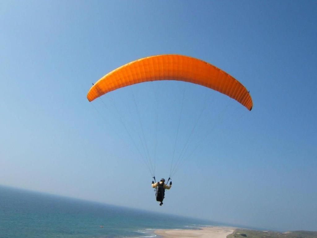 Una persona practicando parapente sobre un paisaje costero con un canopy naranja.