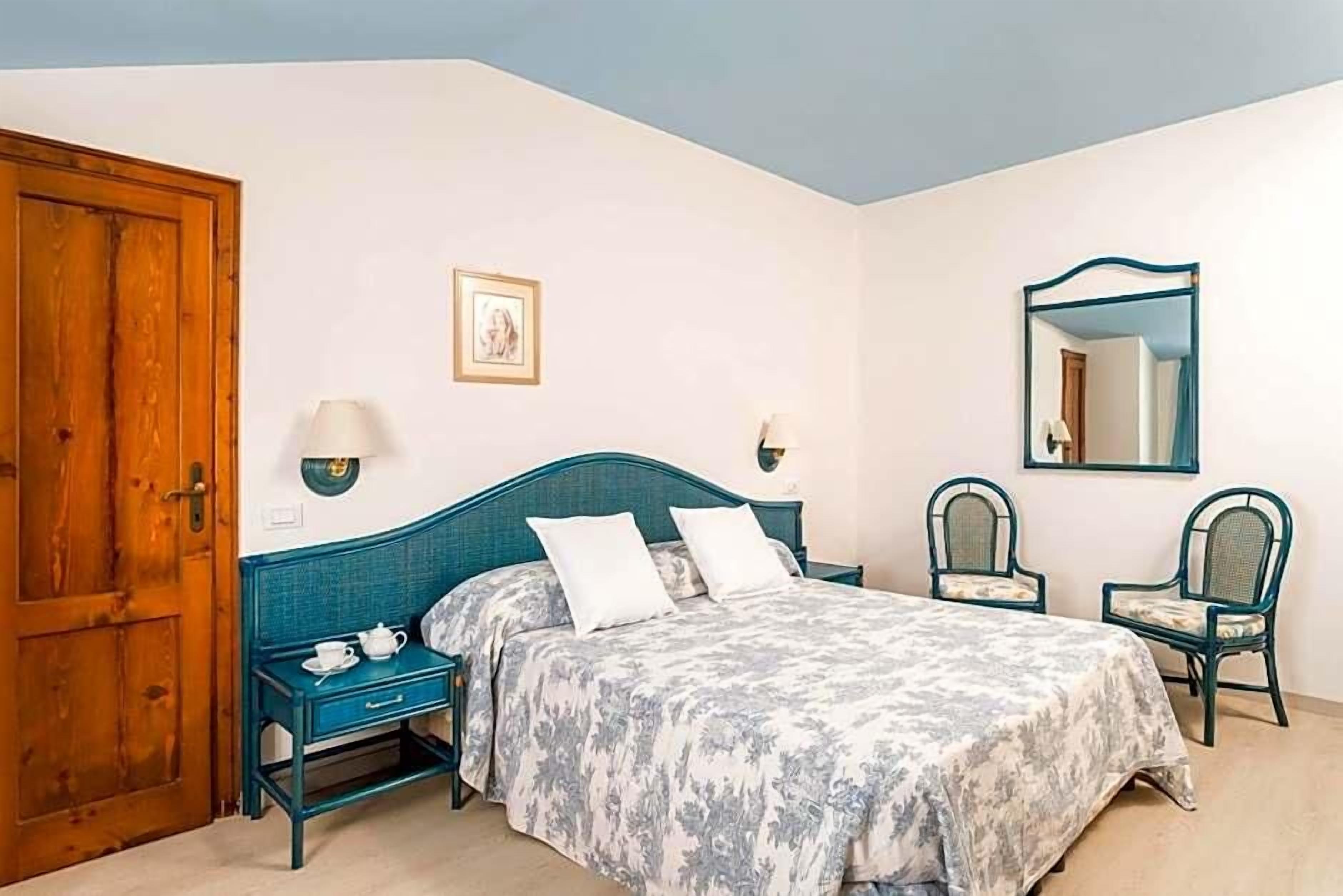 Un dormitorio con cama, mesitas de noche, una silla, una puerta de madera y un espejo en la pared.