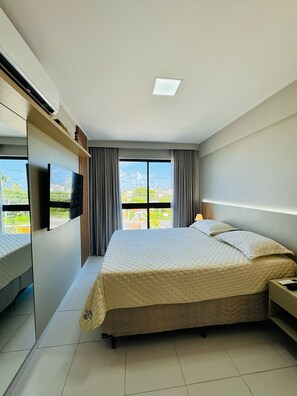 A hotel room with a bed, a TV, a window with curtains, and a view of the outdoors. - 2-bedroom apartment in the Malia Beach Resort in Muro Alto, Porto de Galinhas (Ipojuca)
