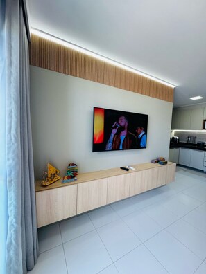A modern living room with a flat-screen TV mounted on the wall, a wooden entertainment unit, and a kitchen area in the background. - 2-bedroom apartment in the Malia Beach Resort in Muro Alto, Porto de Galinhas (Ipojuca)