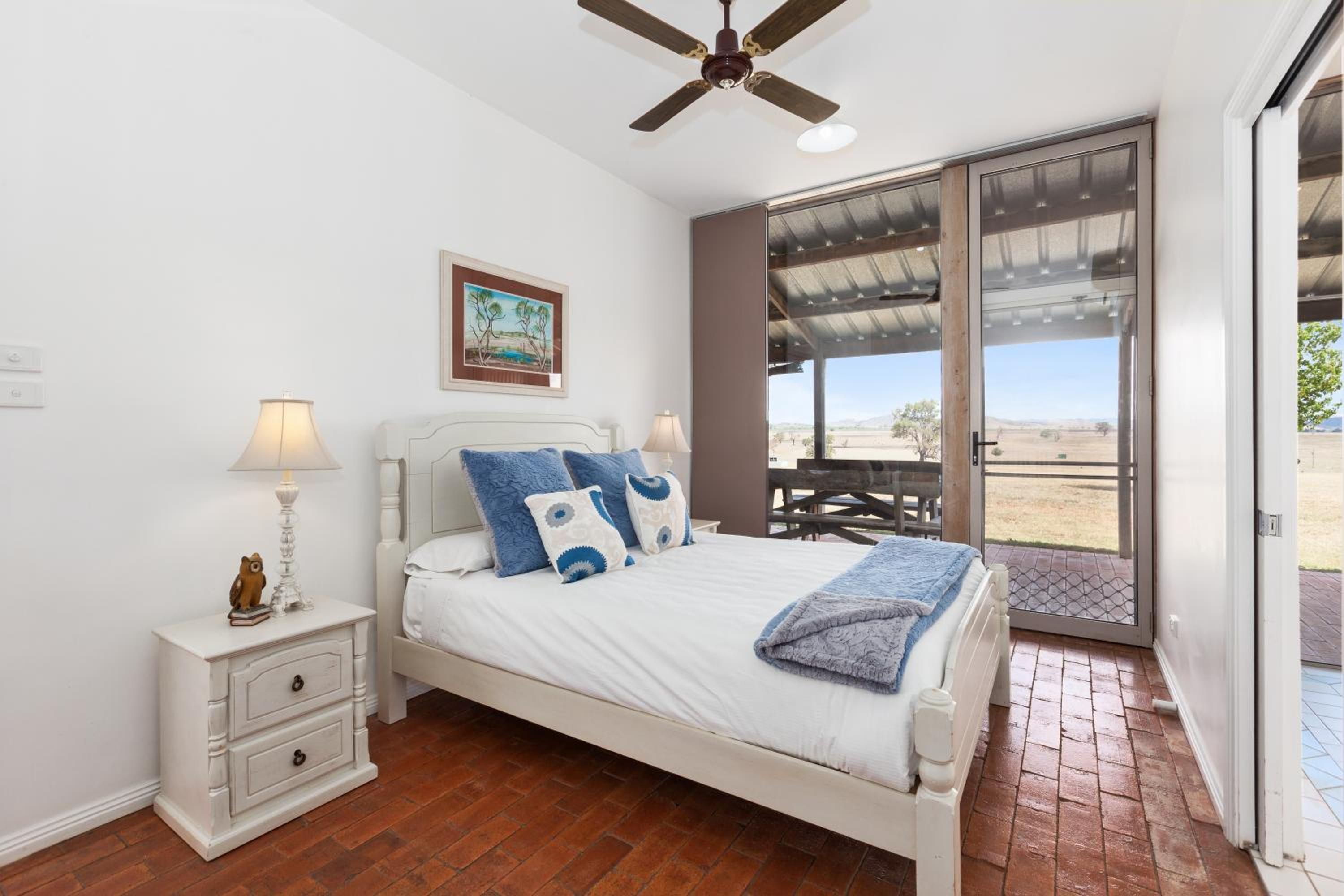 A bedroom with a bed, bedside tables, a ceiling fan, a painting, and a view through a glass door.