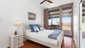 A bedroom with a bed, bedside tables, a ceiling fan, a painting, and a view through a glass door.