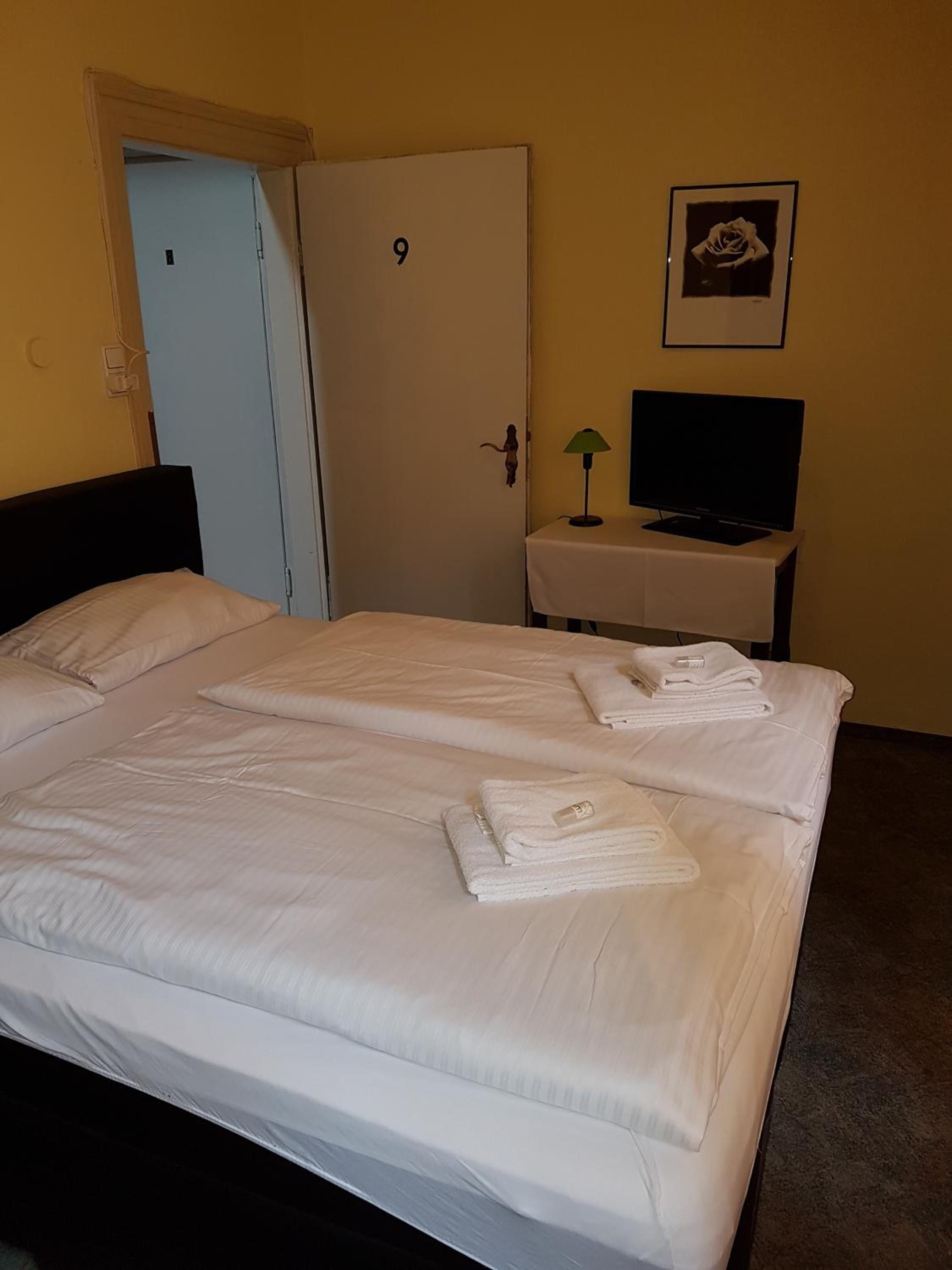 A hotel room with a bed, a television on a stand, a door with a number, and a framed picture on the wall.