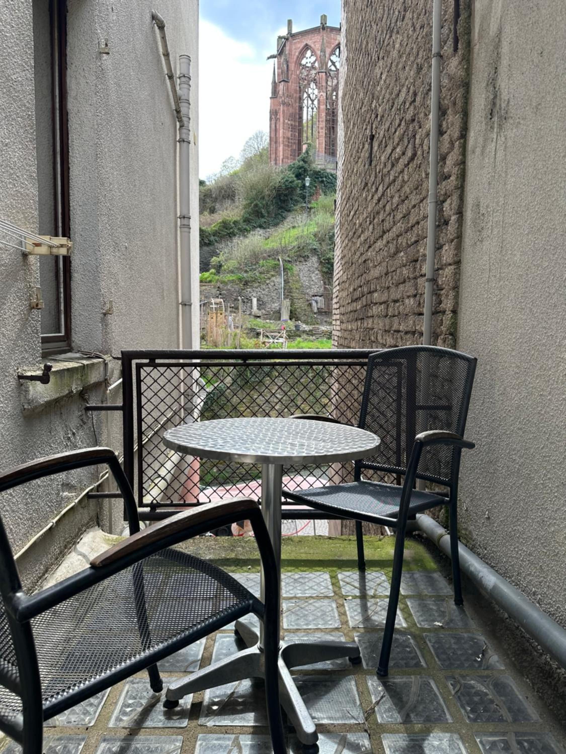 A small outdoor patio with a round metal table and two chairs, a view of a red brick building in the background, and a paved ground.