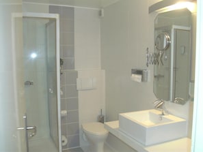 Casa de banho com cabine de duche em vidro, sanita, lavatório com espelho e dispensador de toalhas.