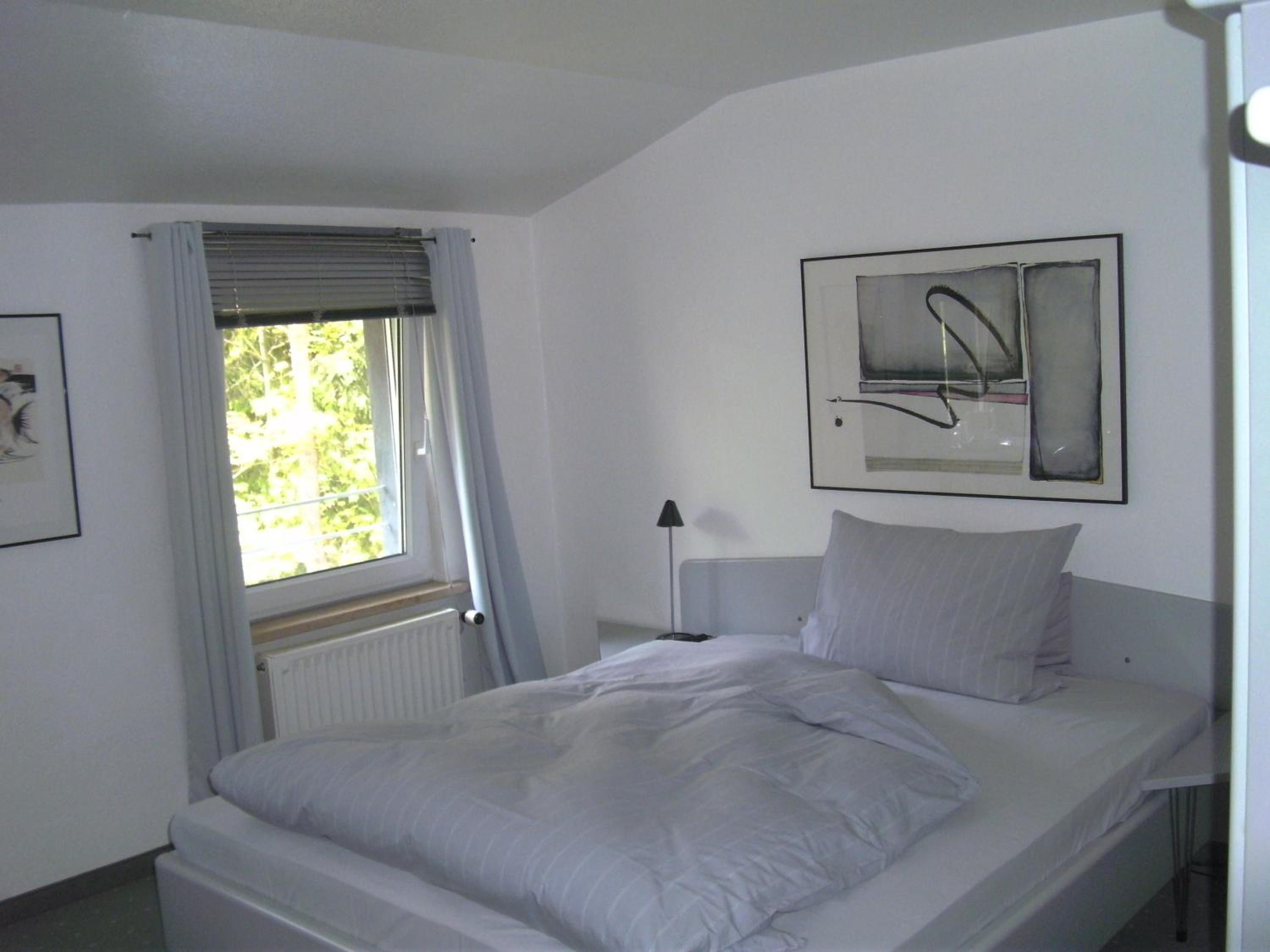 Ein Schlafzimmer mit einem Bett, einem Fenster mit Vorhängen, einem Heizkörper, einer Nachttischlampe und einem gerahmten abstrakten Kunstwerk an der Wand.