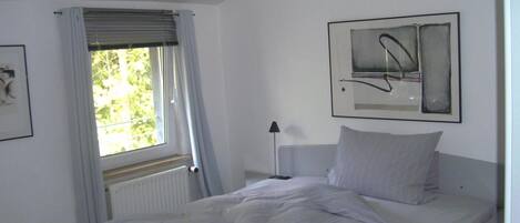 Un dormitorio con cama, ventana con cortinas, radiador, lámpara de noche y una obra de arte abstracta enmarcada en la pared.