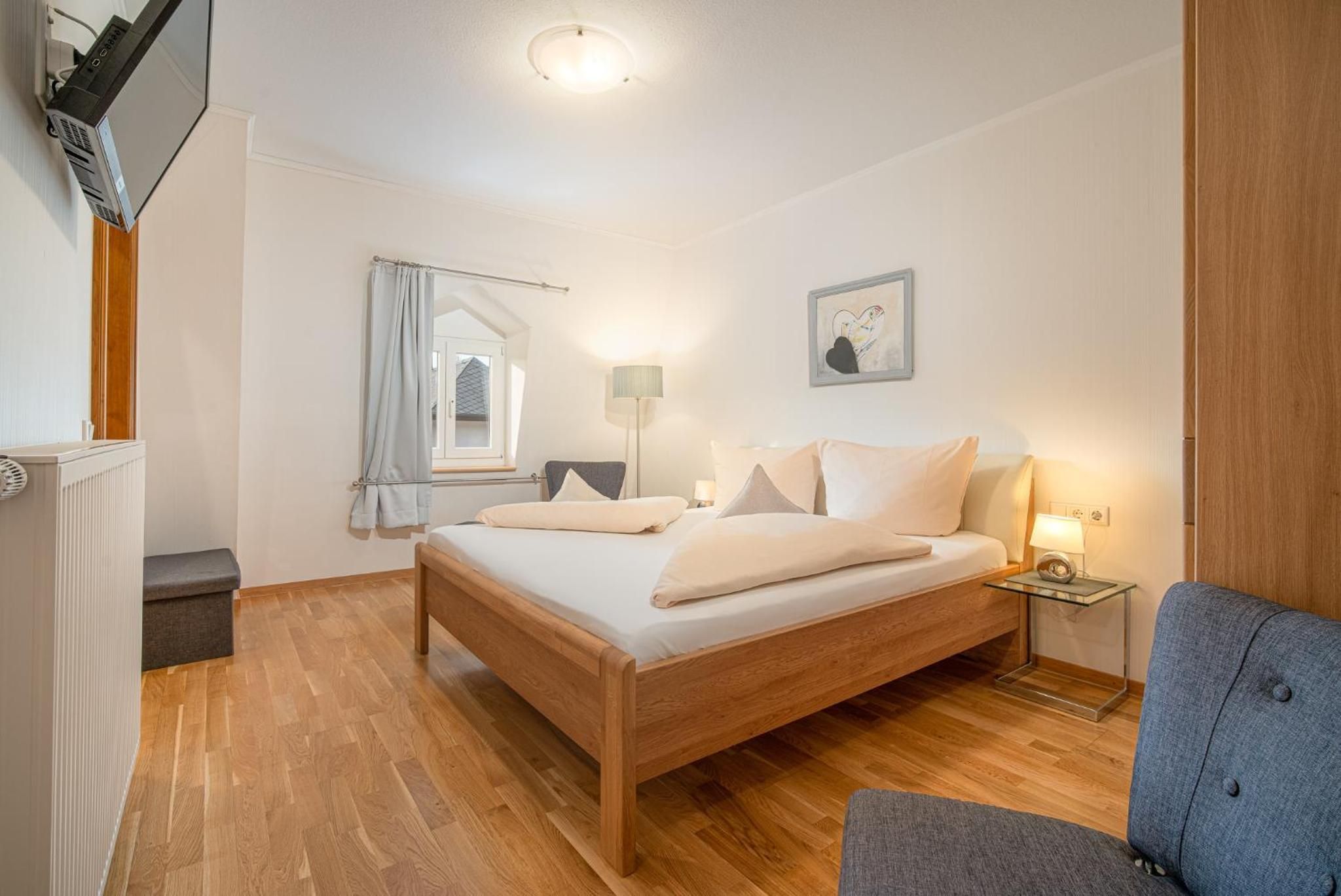 Une chambre à coucher avec un lit en bois, une table de chevet avec une lampe, une fenêtre avec des rideaux et un tableau accroché au mur.