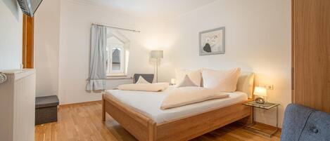A bedroom with a wooden bed, a bedside table with a lamp, a window with curtains, and a wall-mounted picture.