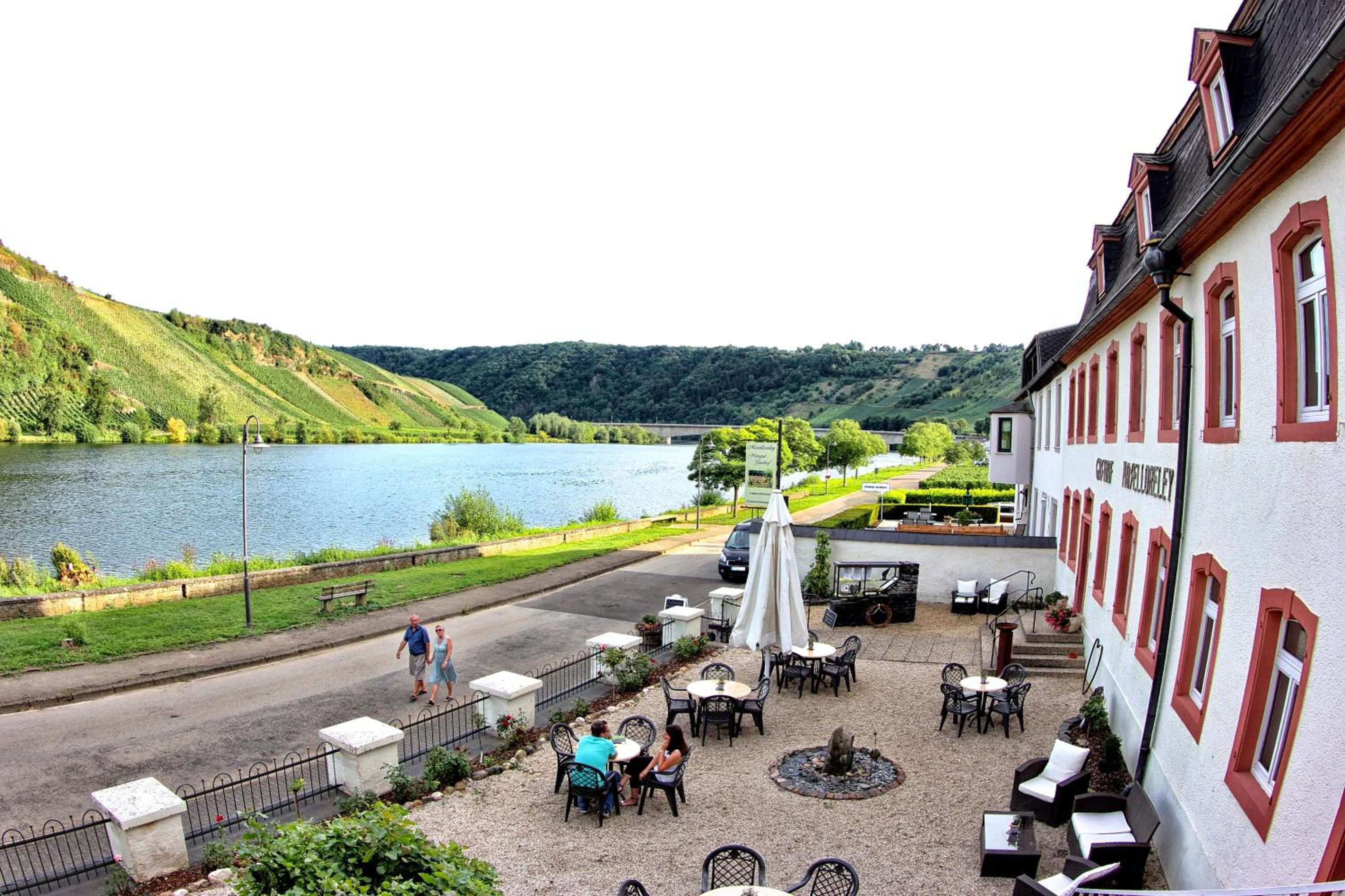 A riverside outdoor seating area with tables and chairs, a fire pit, and a view of a lake and hills.