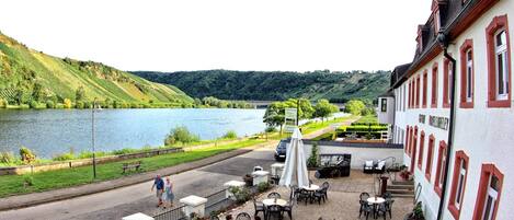 A riverside outdoor seating area with tables and chairs, a fire pit, and a view of a lake and hills.