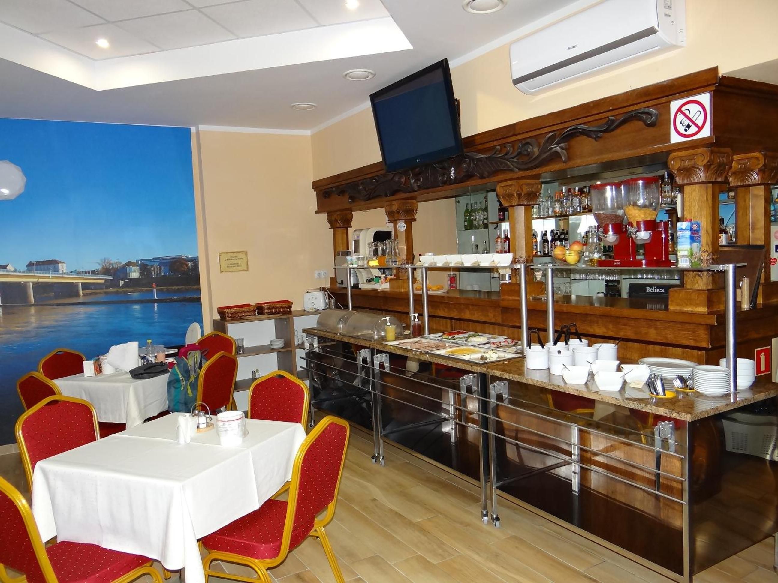 A restaurant with a view of a river, tables with white tablecloths, red chairs, and a bar area with various bottles and glasses.