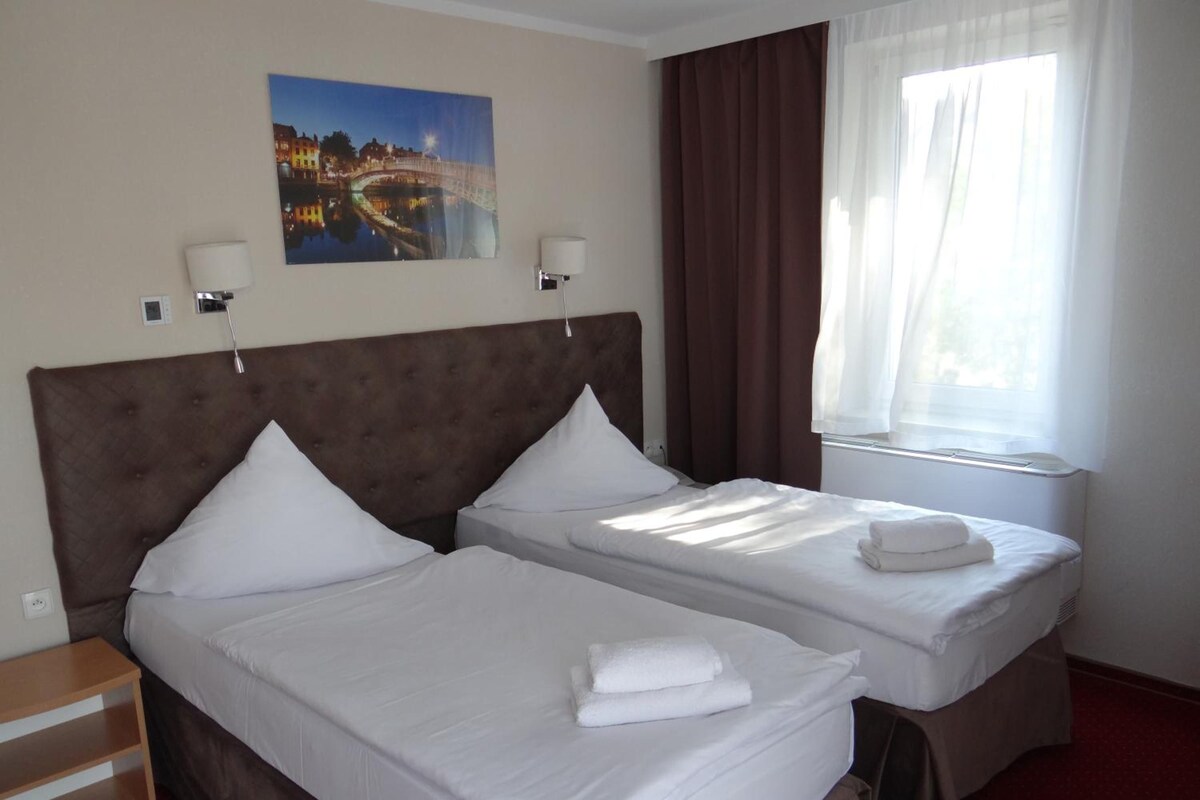A hotel room with two beds, a painting on the wall, a window with curtains, and a bedside table.