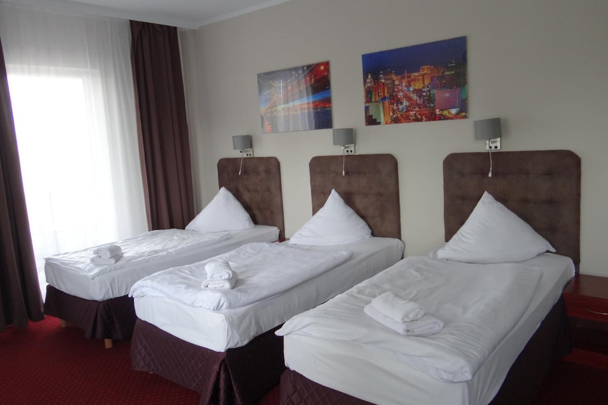A hotel room with two single beds, a large window with curtains, and two wall-mounted lamps.