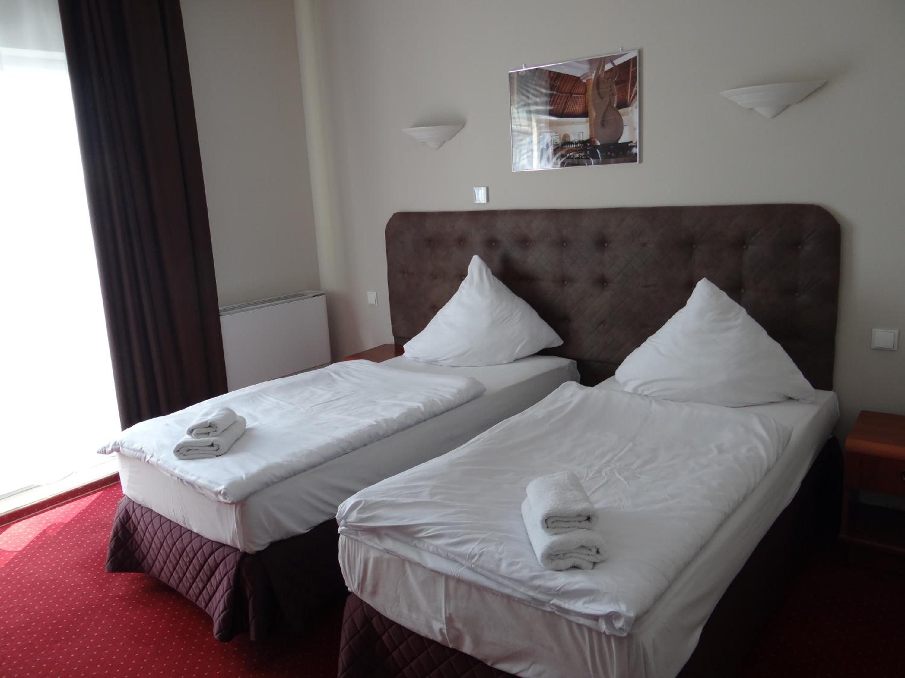 A hotel room with two single beds, each with white linens and towels, a tufted headboard, and a wall-mounted light fixture.