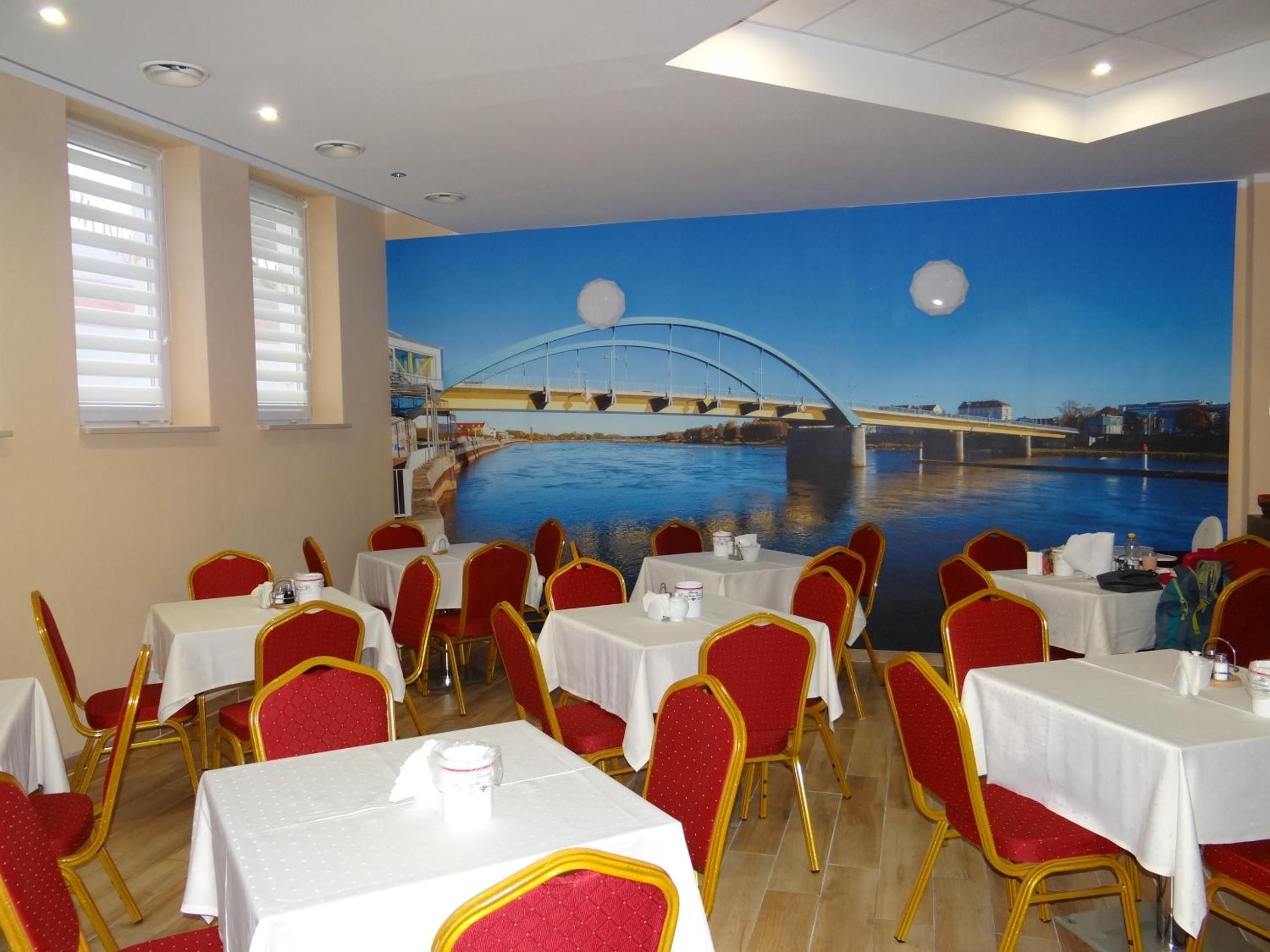 A restaurant with tables and chairs, a mural of a bridge over water, and windows with blinds.