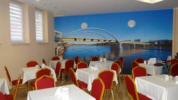 A restaurant with tables and chairs, a mural of a bridge over water, and windows with blinds.