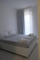 Un dormitorio moderno con una cama grande, ropa de cama blanca y vistas al exterior a través de cortinas traslúcidas.