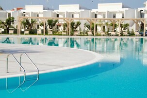 Una piscina cristalina con cubierta de madera y sillas de descanso, rodeada de edificios blancos con balcones.