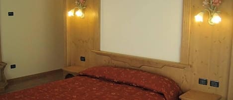 Un dormitorio con cabecera de madera, colcha roja, dos lámparas de pared y una mesita de noche con cajón.