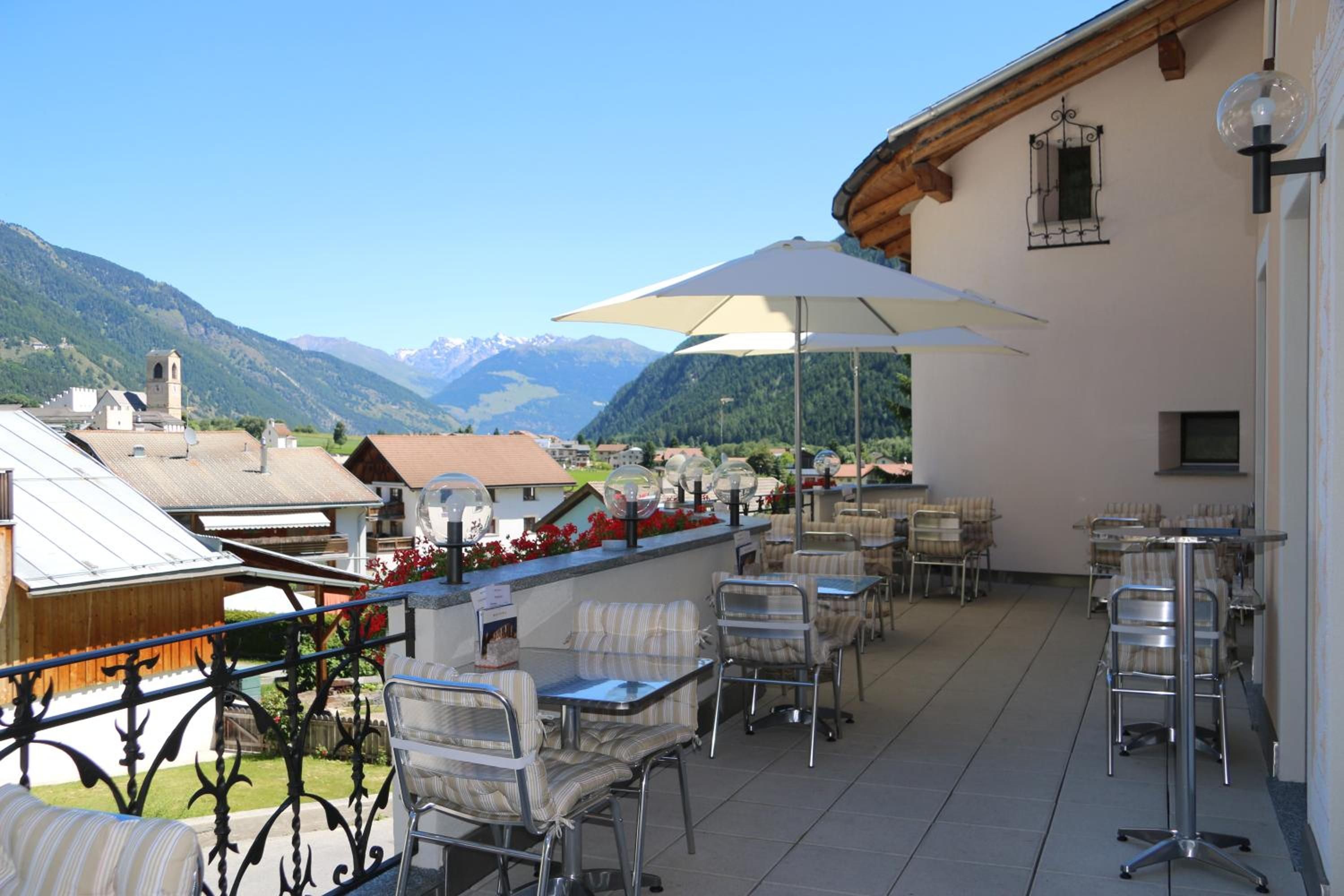 Eine Terrasse im Freien mit Tischen und Stühlen, einem großen Sonnenschirm und Blick auf Berge.