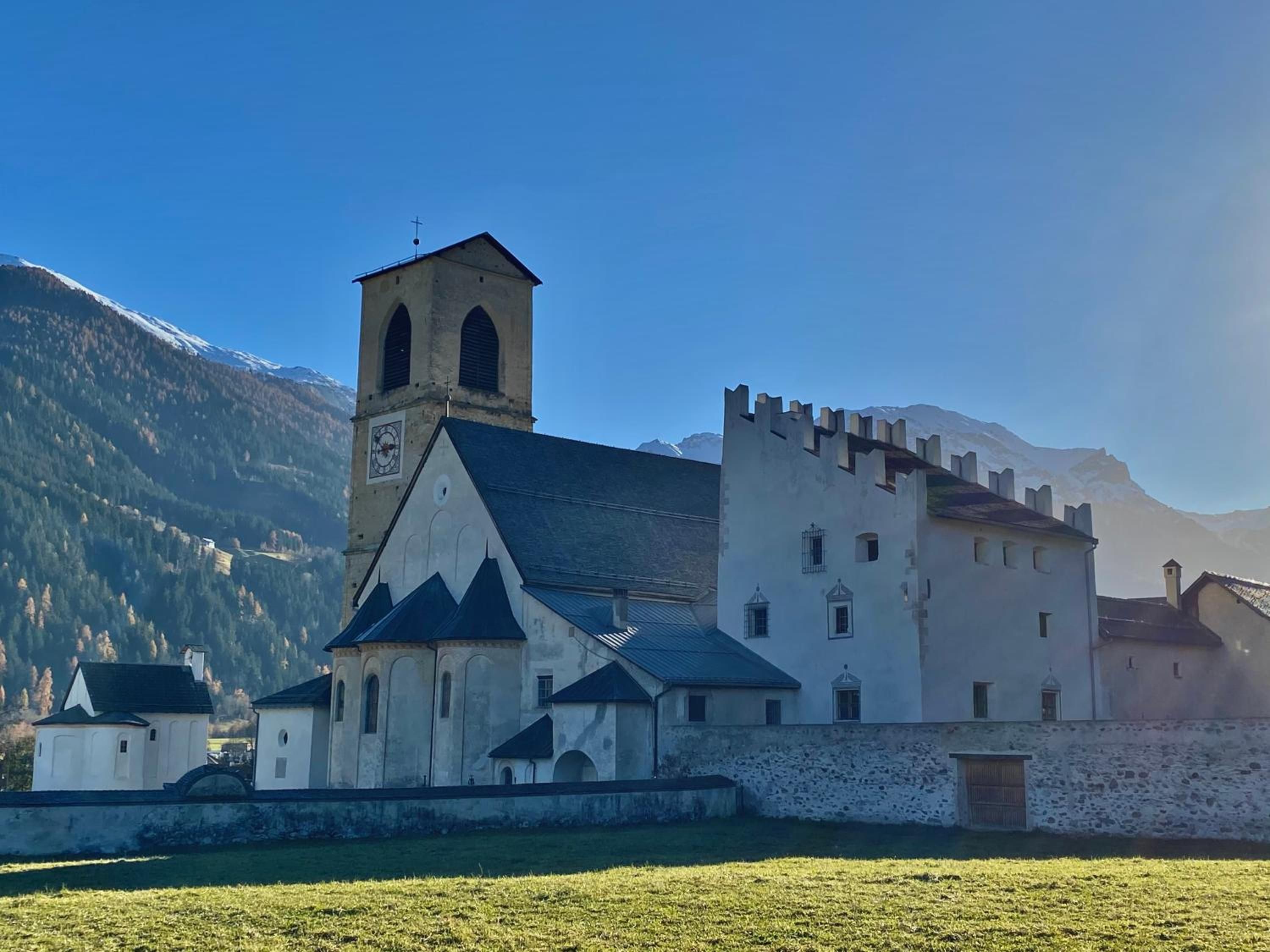 Un edificio storico con un campanile e una struttura fortificata, circondato da un prato erboso e montagne.