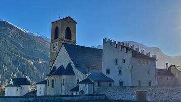 Un edificio storico con un campanile e una struttura fortificata, circondato da un prato erboso e montagne.