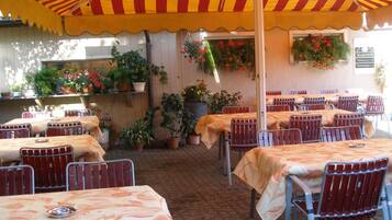 An outdoor dining area with tables and chairs under a striped canopy, surrounded by potted plants and a brick wall.