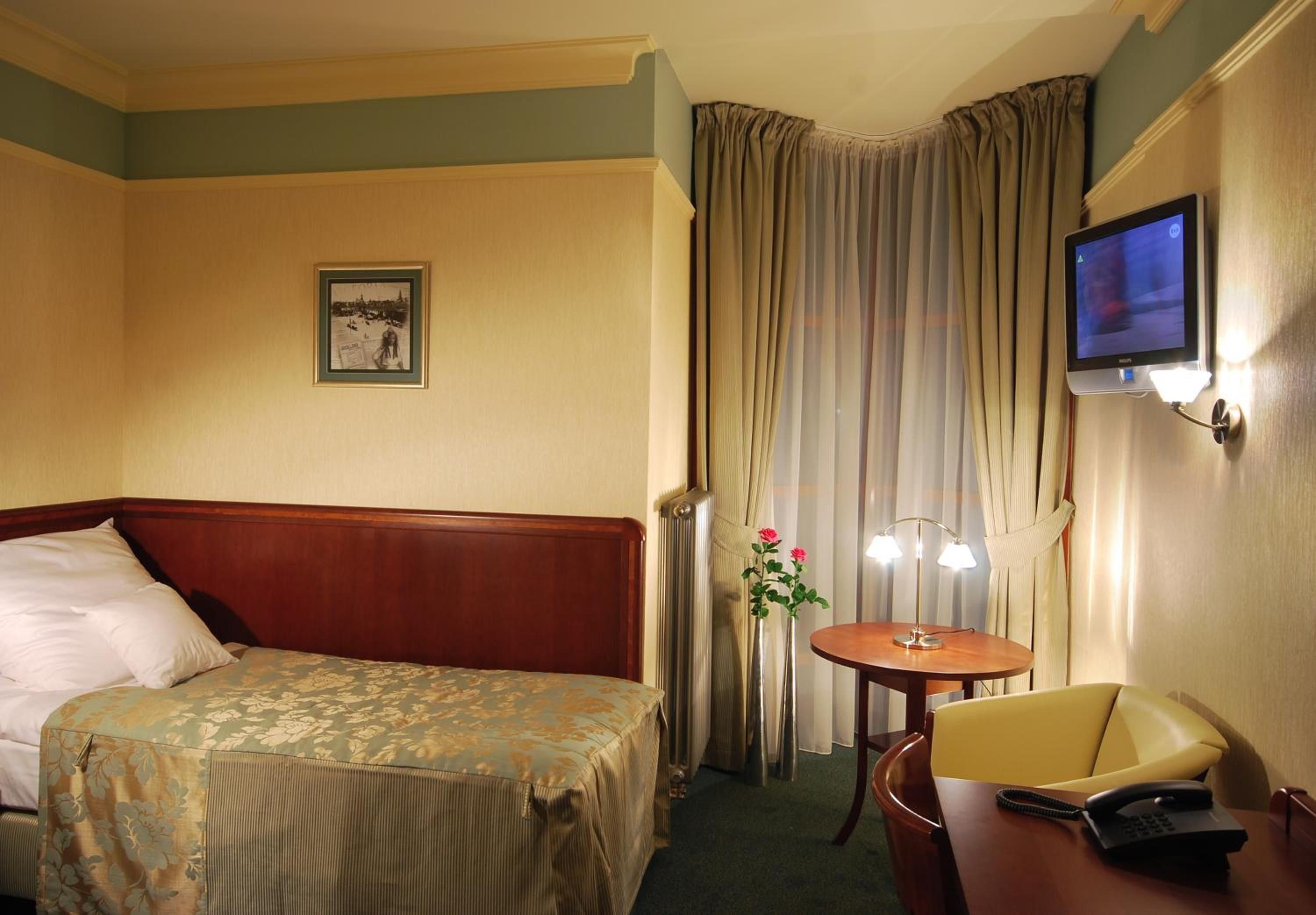 A hotel room with a bed, a desk with a chair, a television, and a window with curtains.