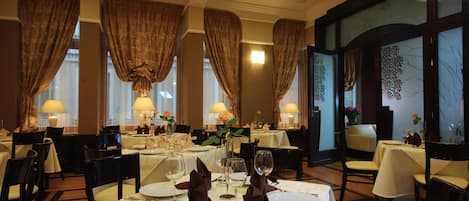 A restaurant with multiple tables set for dining, featuring white tablecloths and gold-toned cutlery.