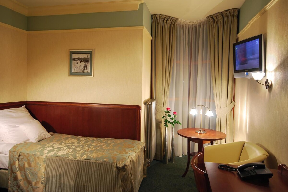 A hotel room with a bed, a desk with a chair, a television, and a window with curtains.