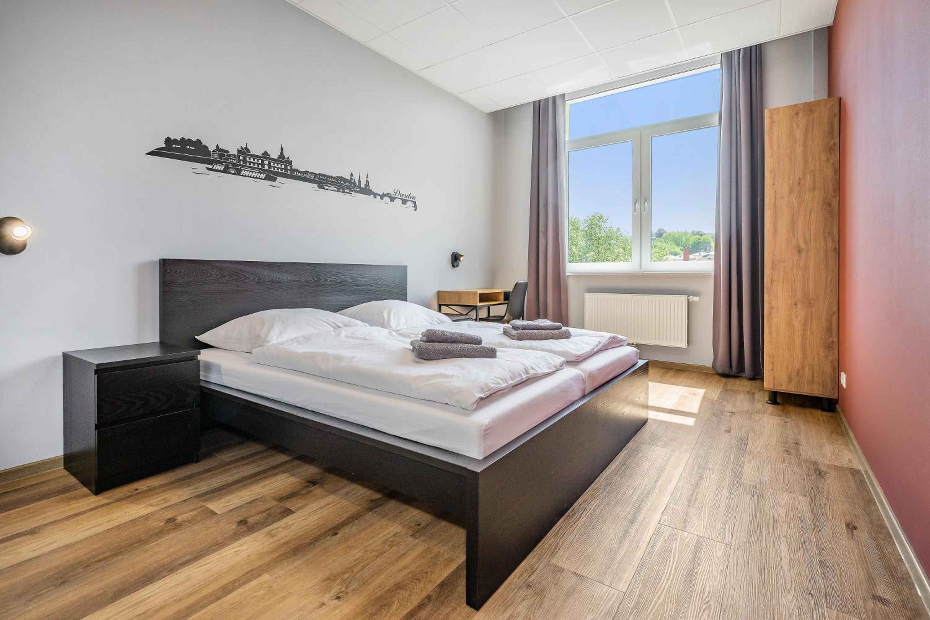 A modern hotel room with a large bed, a nightstand, a wooden floor, a cityscape wall art, a window with curtains, and a radiator.