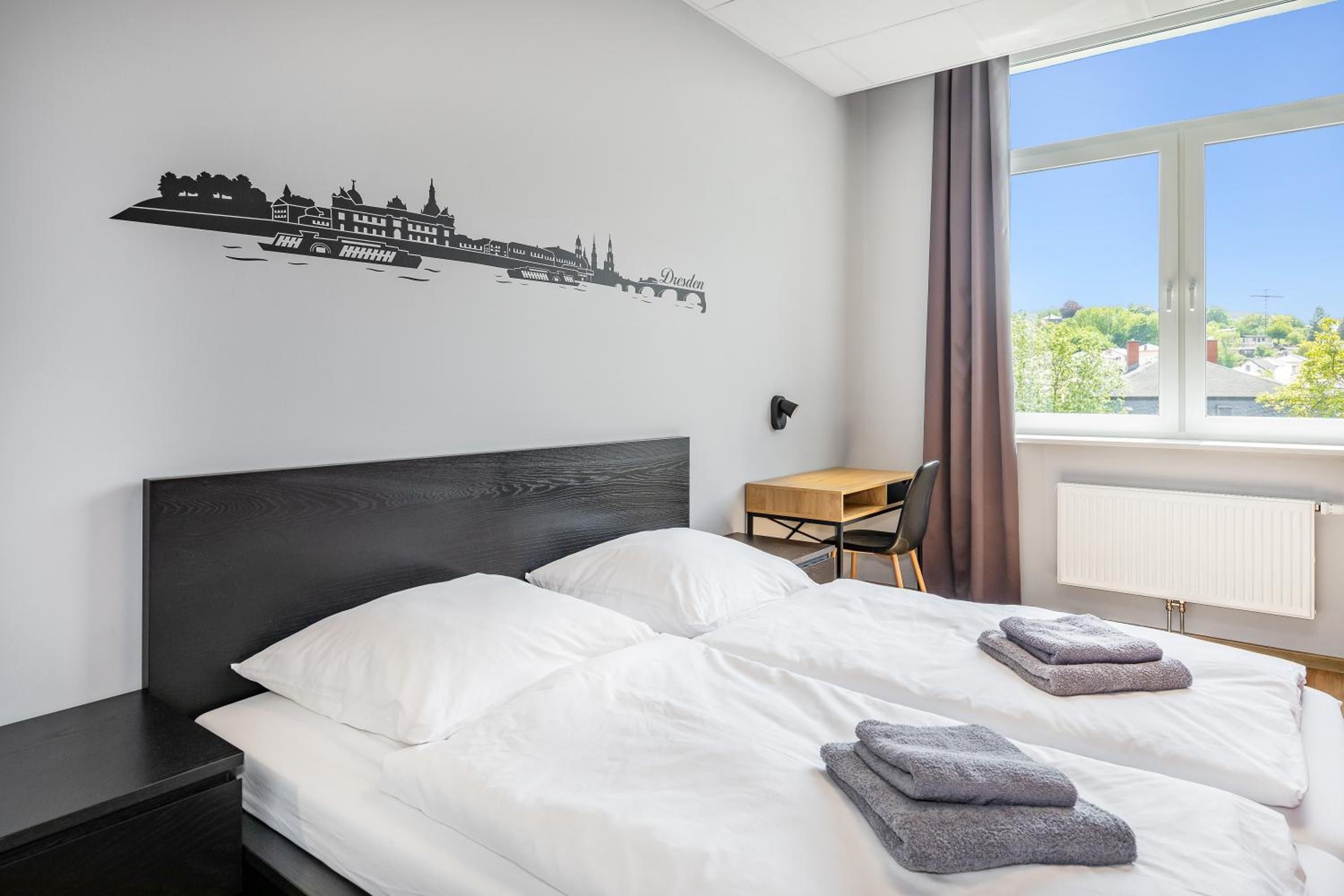 A hotel room with a bed, a desk, a chair, and a window with a view of buildings.