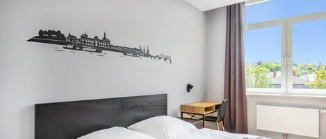 A hotel room with a bed, a desk, a chair, and a window with a view of buildings.