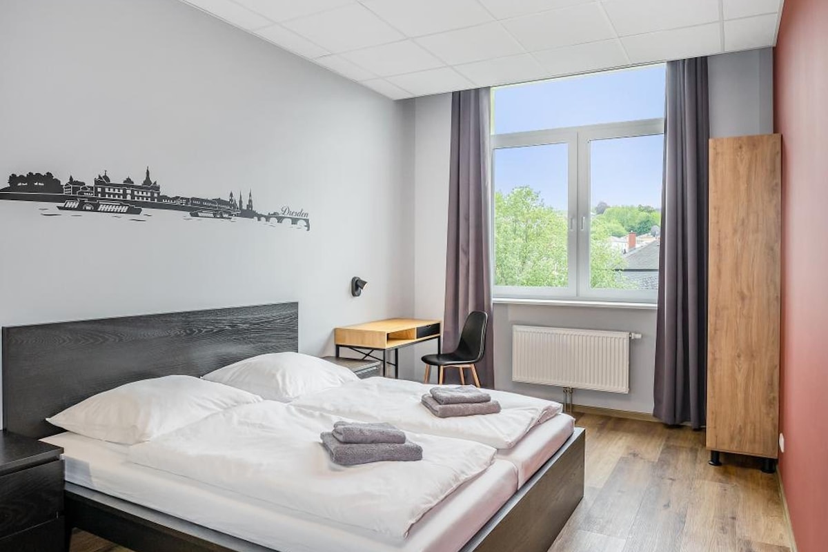 Habitación de hotel moderna con una cama grande, un escritorio, una silla y vistas al exterior a través de una ventana.