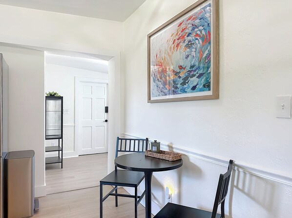 Un coin repas avec une table ronde, deux chaises, une poubelle noire, un luminaire fixé au mur et une porte donnant sur une autre pièce.