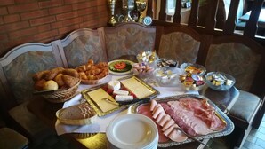 Una mesa dispuesta con diversos alimentos, entre ellos pasteles, quesos y carnes, en una habitación con mobiliario estampado y una pared de ladrillo.