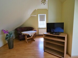 A living room with a television on a stand, a sofa, a small table with a tablecloth, and a potted plant. - Gasthof Erfurth (Sehnde)