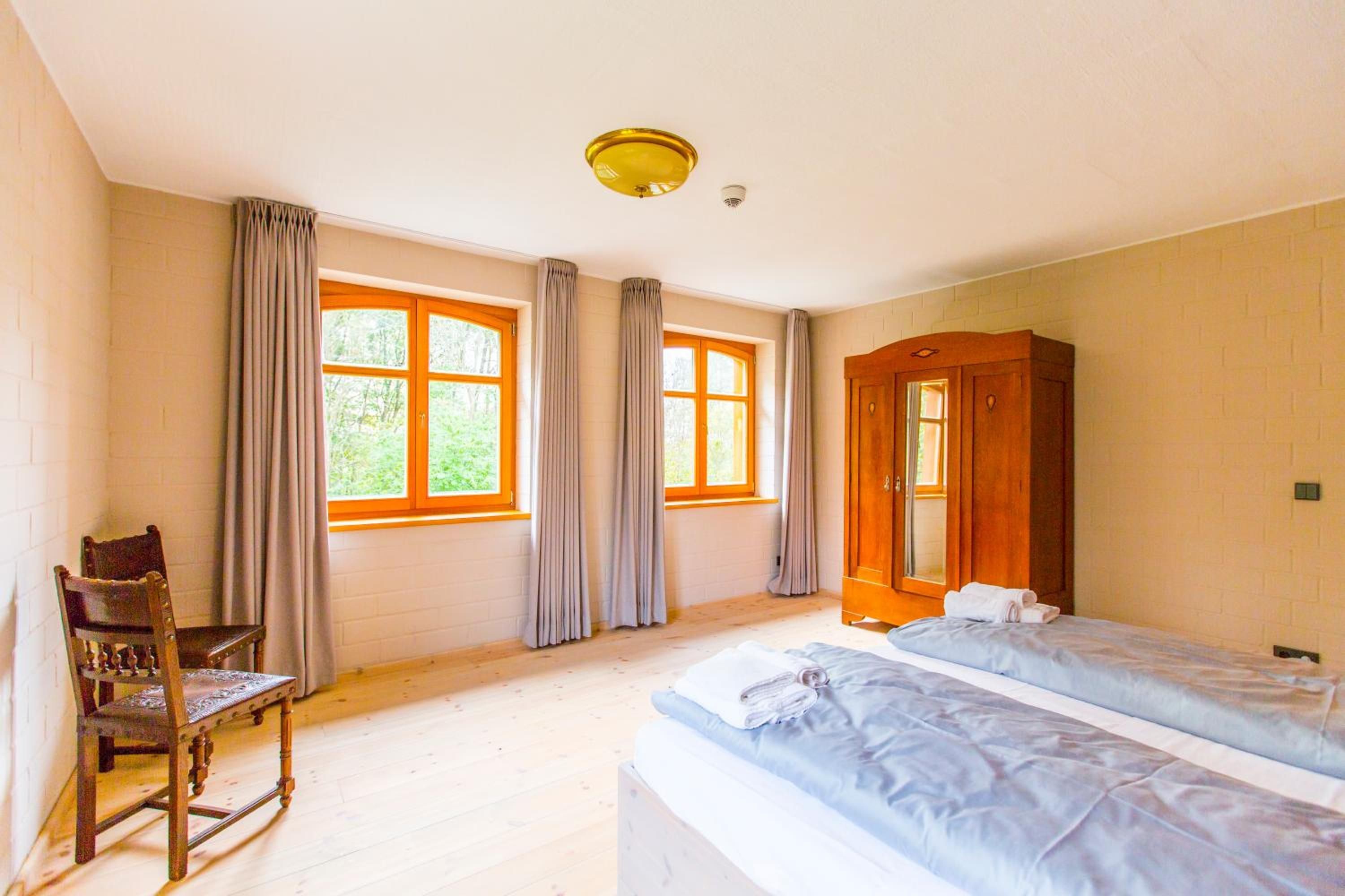 A room with two beds, a wooden wardrobe, a chair, and large windows with curtains.